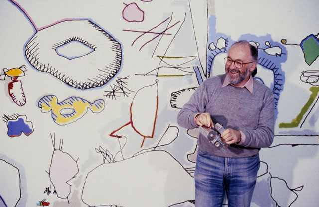 Harold Cohen holding a Turtle Robot at the San Francisco Museum of Modern Art, 1979 with a computer-generated, enlarged and hand-coloured temporary mural in the background. Courtesy Harold Cohen’s archive.