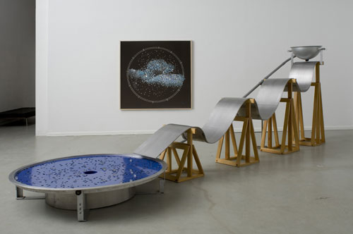 Alice Aycock. Some Night Action, 1993. Aluminum, wood, marbles, motorized parts, an undulating ramp down which marbles flow into a spinning star system; 6 h x 17 l x 5 w ft. Clear glass marbles are ejected from a pipe into a bowl and flow down an undulating ramp into a spinning circular dish with slots representing the constellations. The marbles remain in their respective places until they are knocked out of position by other shooting marbles. The marbles fall into a center void, only to be recycled again. Photograph: Tim Lee