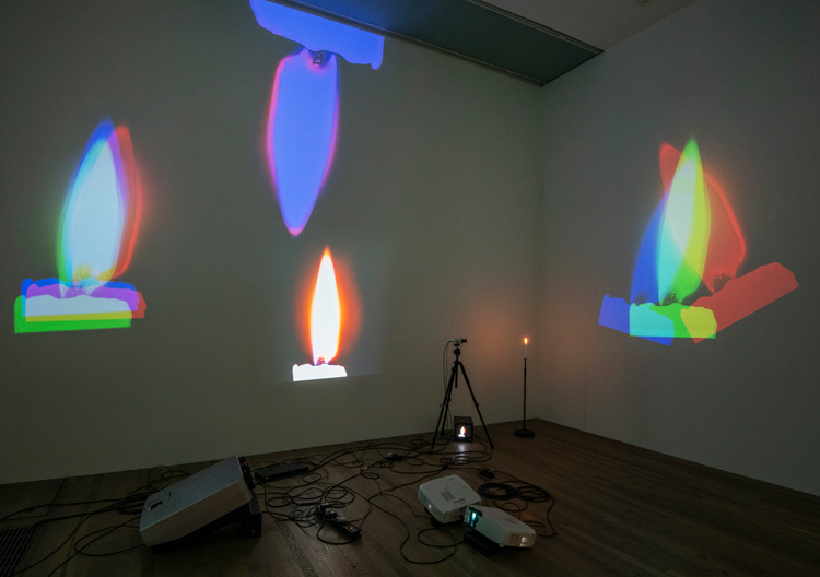 Nam June Paik. One Candle (Candle Projection) 1989. Installation view, Tate Modern 2019. Close-circuit television camera, tripod, candle on custom stand and four to six colour video projectors including modified CRT projectors. Museum für Moderne Kunst, Frankfurt am Main.